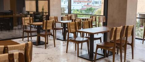 A spacious dining area with wooden tables and chairs, large windows offering a view of buildings outside.