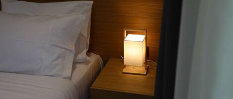 Uma cama bem arrumada com lençóis brancos, cabeceira de madeira e mesa de cabeceira com uma luminária acesa.