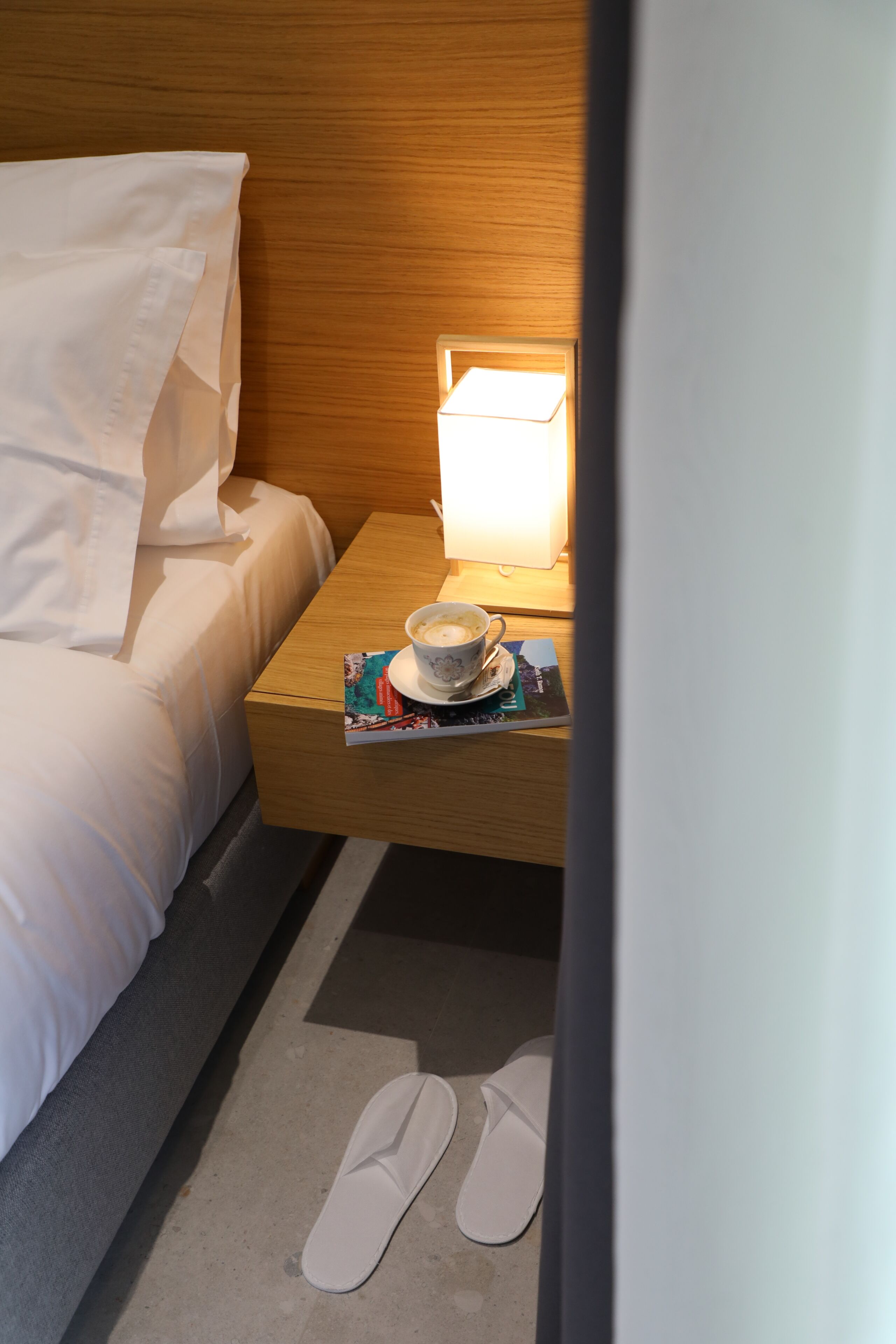 A hotel room with a bed, a nightstand with a lamp, a cup, and a book.