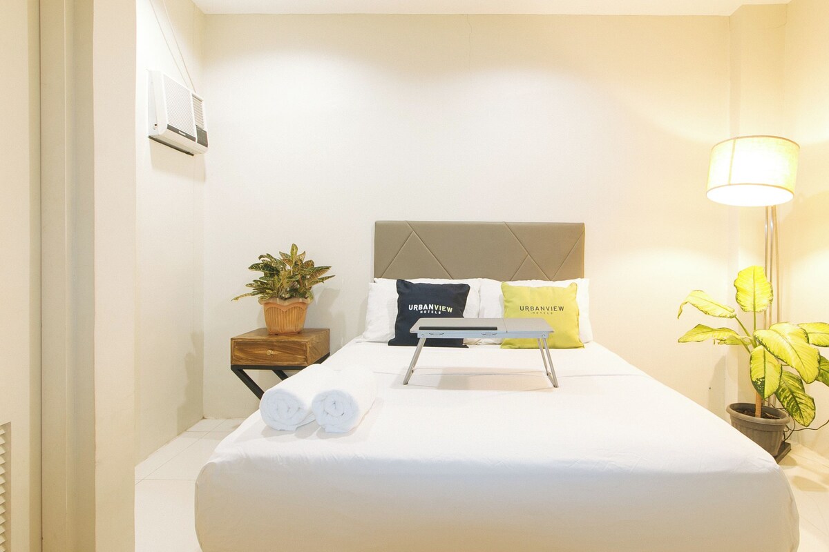 A neatly made bed with two pillows, a bedside table with a plant, and a lamp on the right.