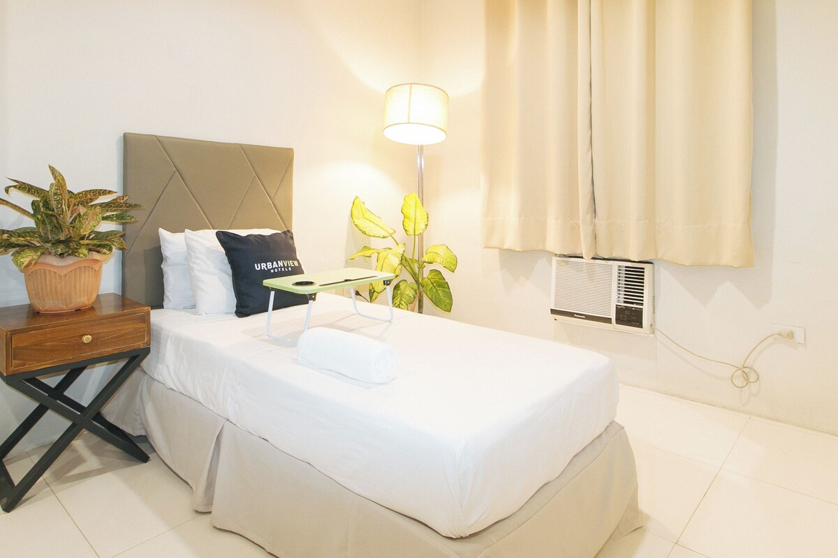 A neatly made bed with a pillow, a bedside table with a plant, a lamp, and a window with curtains.