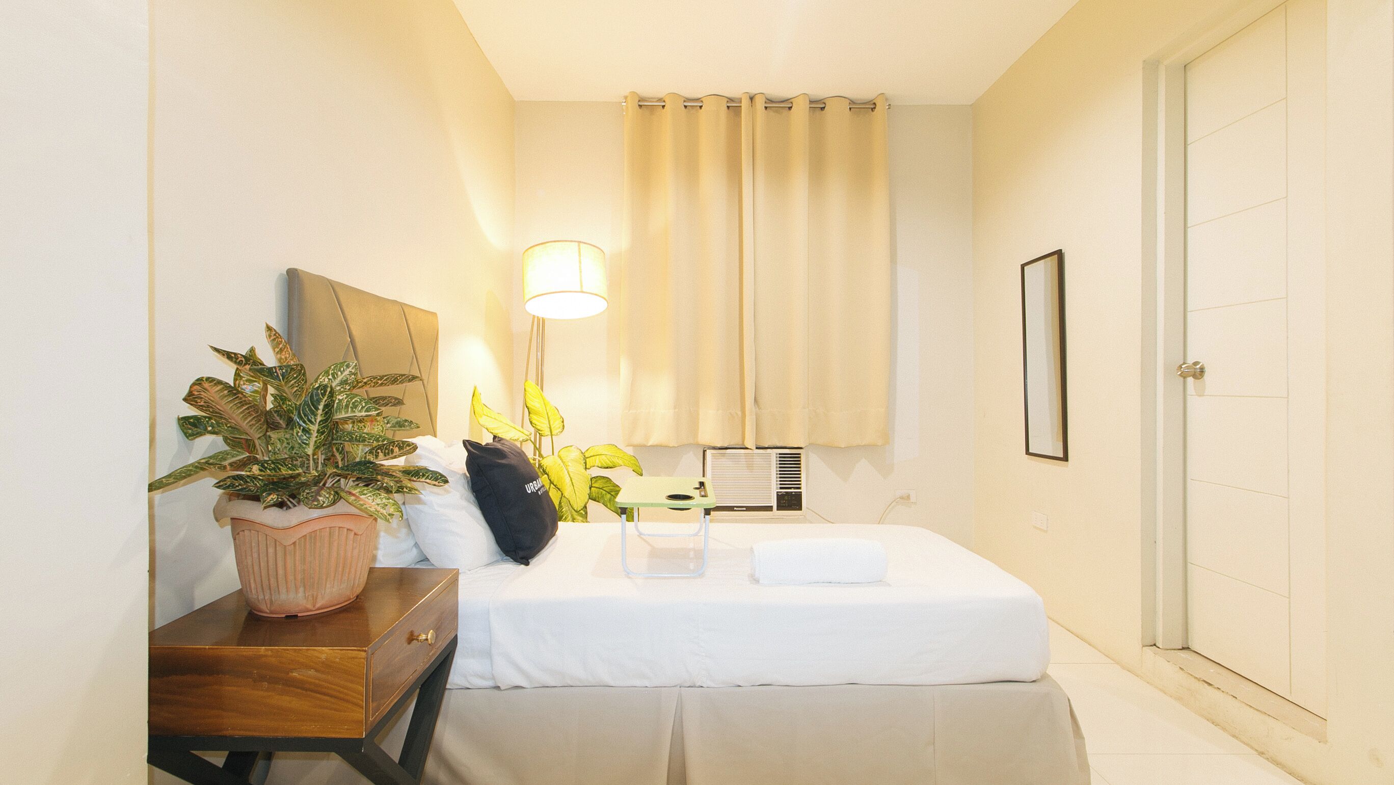 Habitación de hotel ordenada con cama, mesita de noche, plantas en macetas, lámpara y espejo.