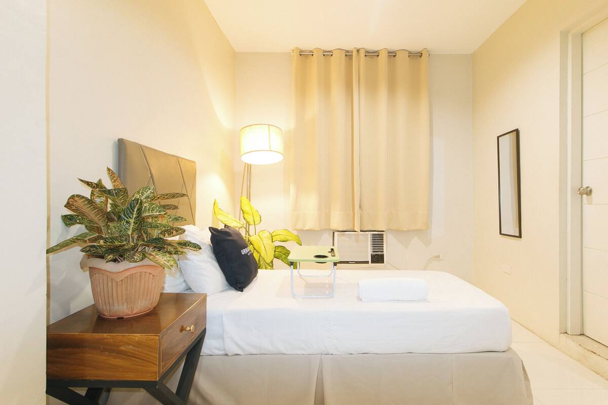 Une chambre d’hôtel bien rangée, avec un lit, une table de chevet, des plantes en pot, une lampe et un miroir.