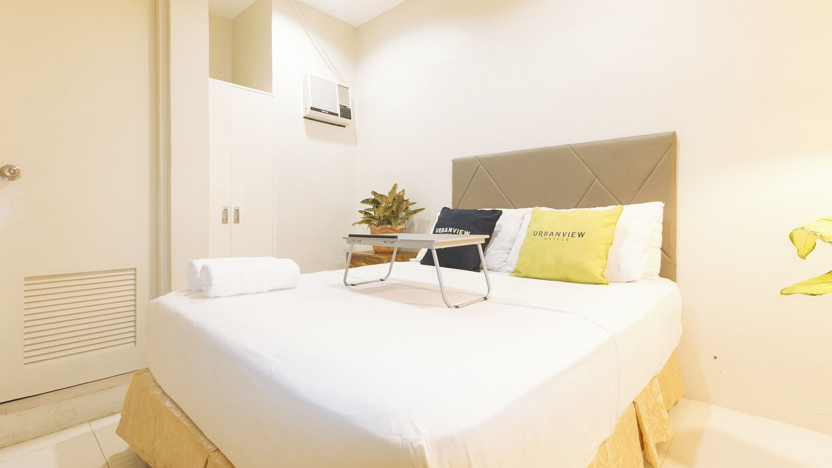 Una habitación de hotel con una cama, una mesita de noche, una planta en maceta y un aire acondicionado de pared.