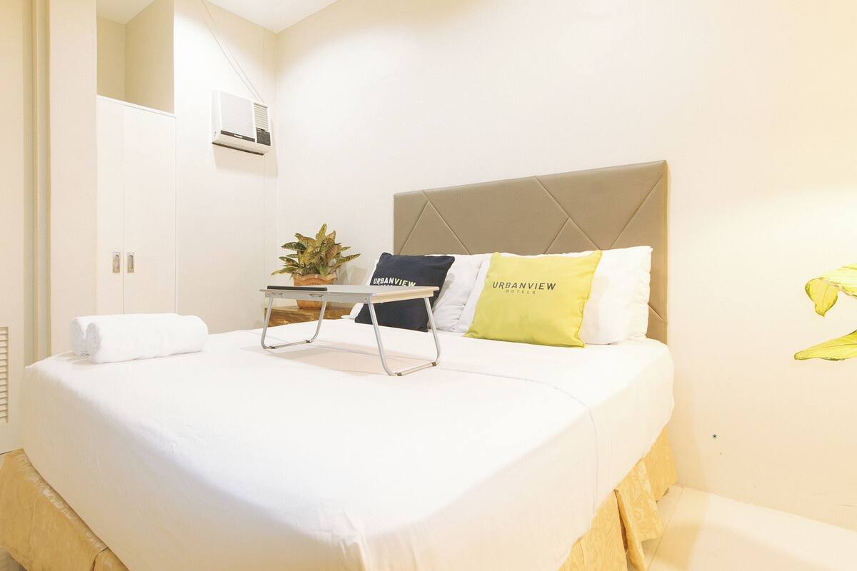 Une chambre d’hôtel avec un lit, une table de chevet, une plante en pot et un climatiseur fixé au mur.
