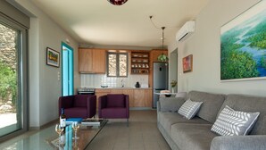 Ein modernes Wohnzimmer mit einem Glastisch, zwei violetten Sesseln, einem grauen Sofa mit gemusterten Kissen und einer Küche im Hintergrund.