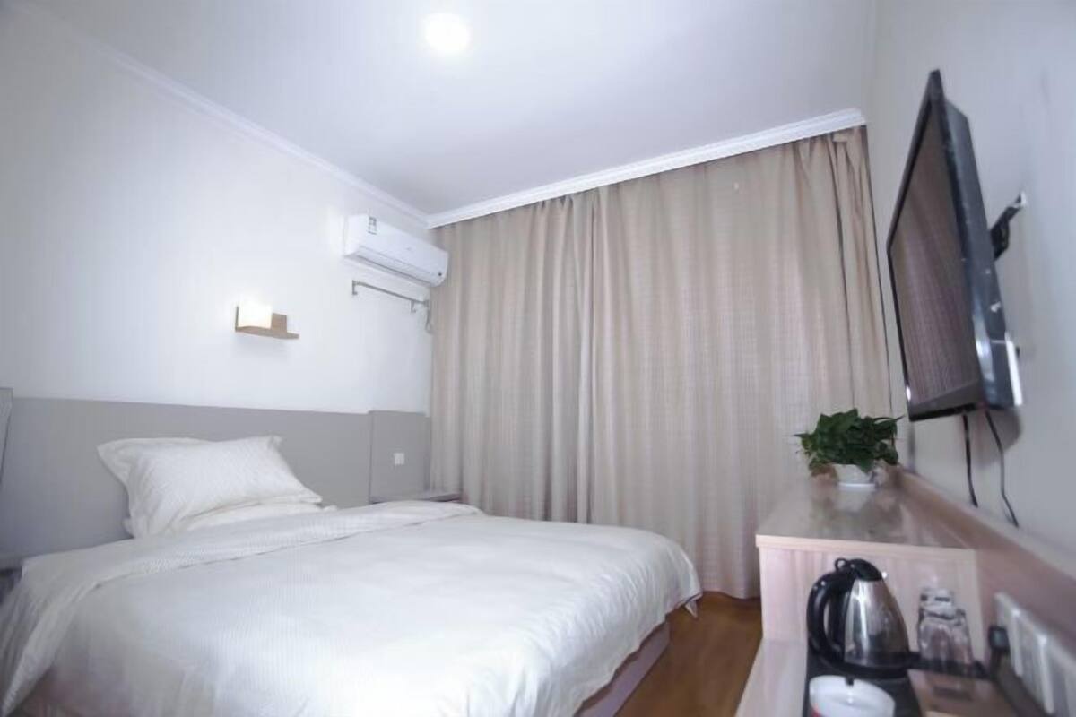 A hotel room with a bed, a TV, an air conditioning unit, a window with curtains, and a small table with a plant and a kettle.