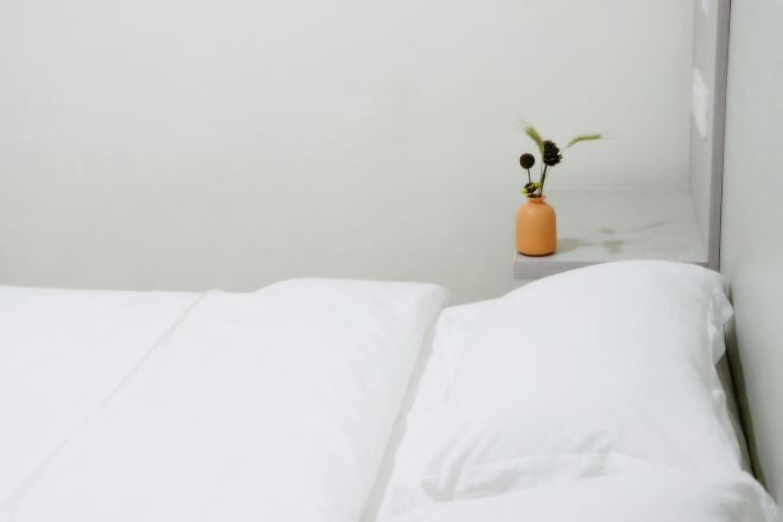 A bed with white sheets, a small orange vase with flowers, and a white wall.