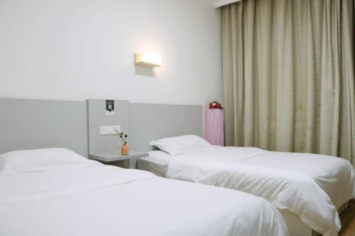 A hotel room with two single beds, a bedside table with a lamp, a wall-mounted light, and a window with curtains.