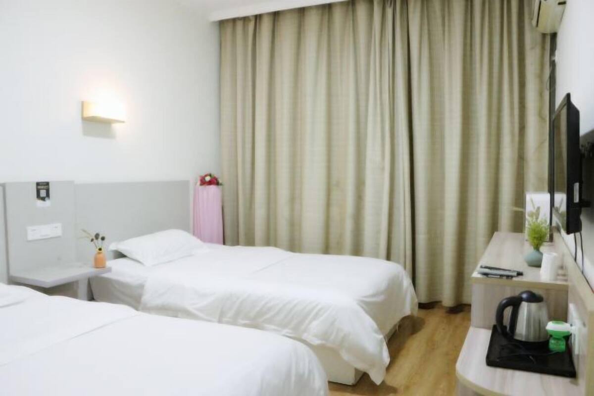 A hotel room with two single beds, a desk with a kettle, a small table with a vase, a mirror, and a TV on a stand.