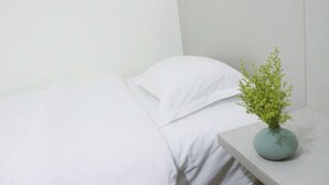 Ein ordentlich bezogenes Bett mit weißen Leinen und einer kleinen, grünen Pflanze in einer runden Vase auf einem Nachttisch.