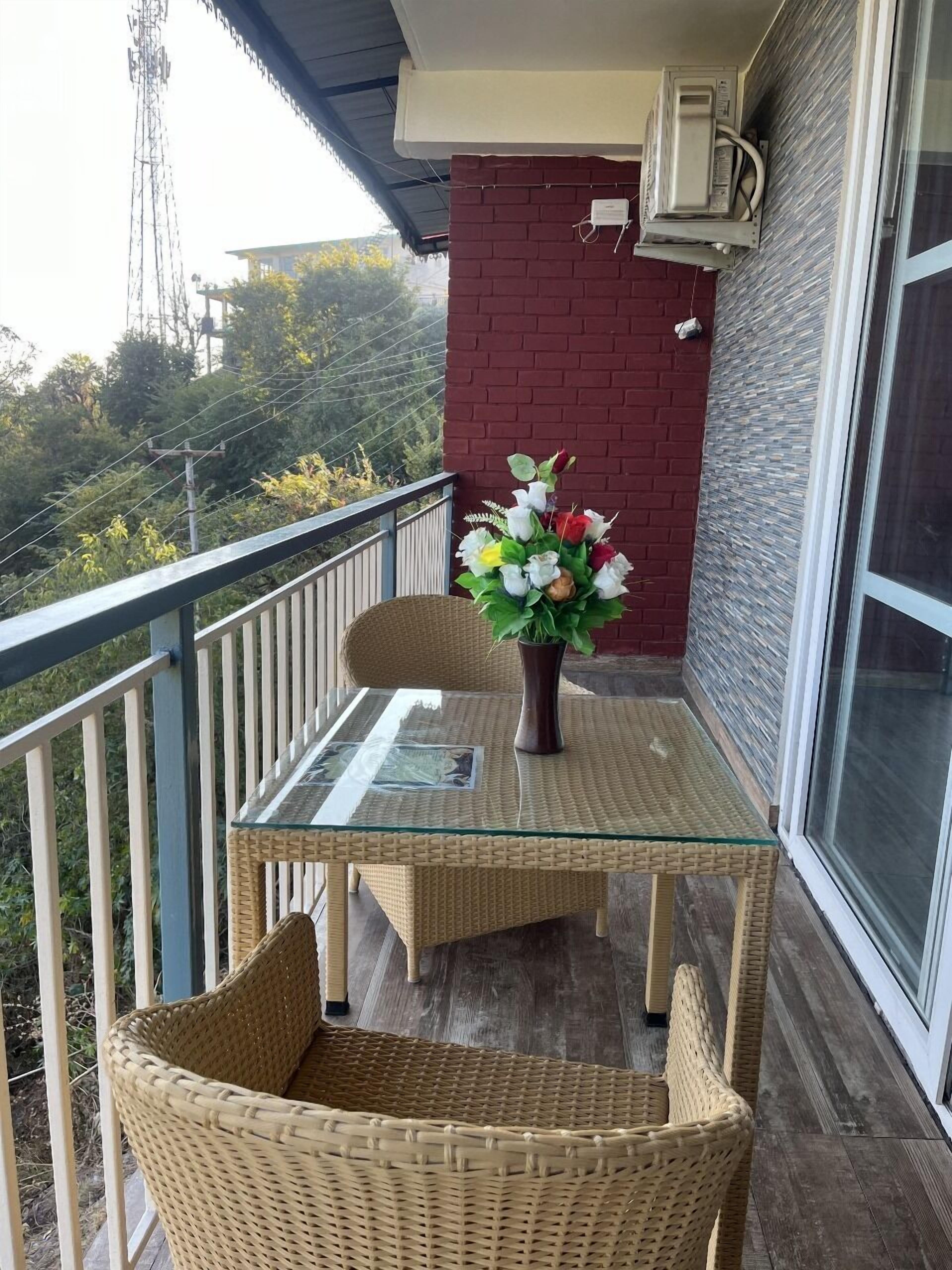 Un balcón con una mesa de vidrio y sillas de mimbre, un jarrón con flores y un muro de ladrillo rojo.