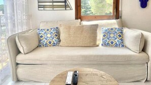 Un sofá color arena con cojines estampados, una mesa de centro de madera con dos mandos a distancia y una ventana con vistas a árboles.