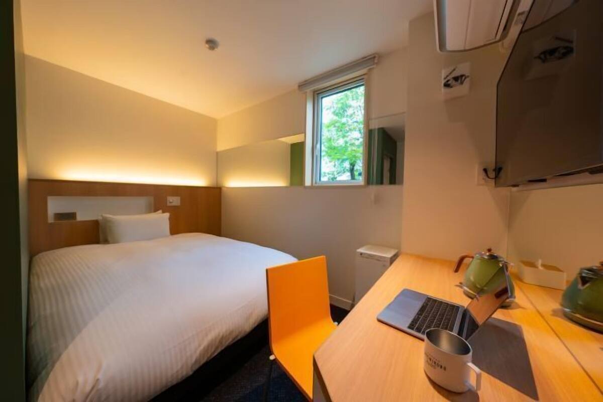 Une chambre d’hôtel avec un lit, un bureau avec un ordinateur portable, une tasse et une fenêtre donnant sur des arbres.