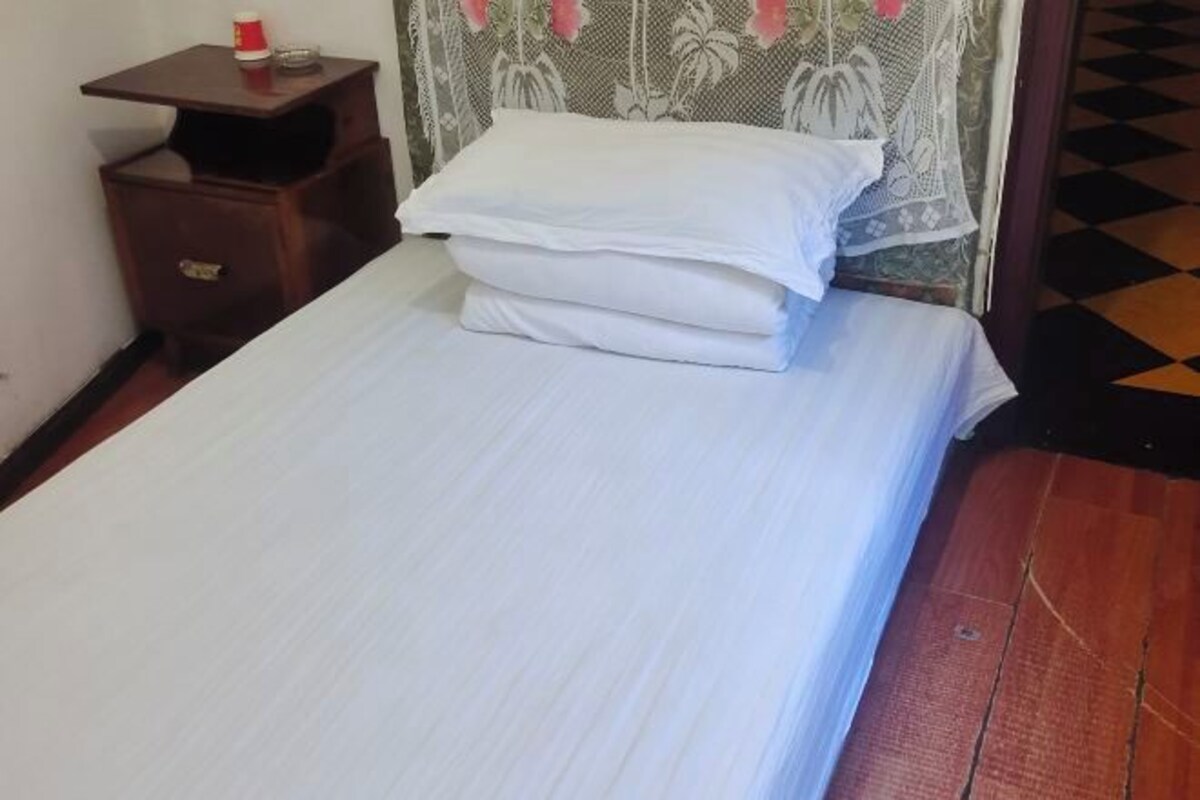 A neatly made bed with white linens, a small wooden nightstand with a bottle and glass, and a patterned curtain.