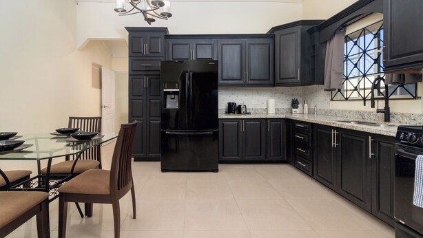 A modern kitchen with dark cabinetry, a black refrigerator, a dining table with chairs, and a window with a geometric design. - 5 Bedroom Haven in Draxhall (Ocho Rios)