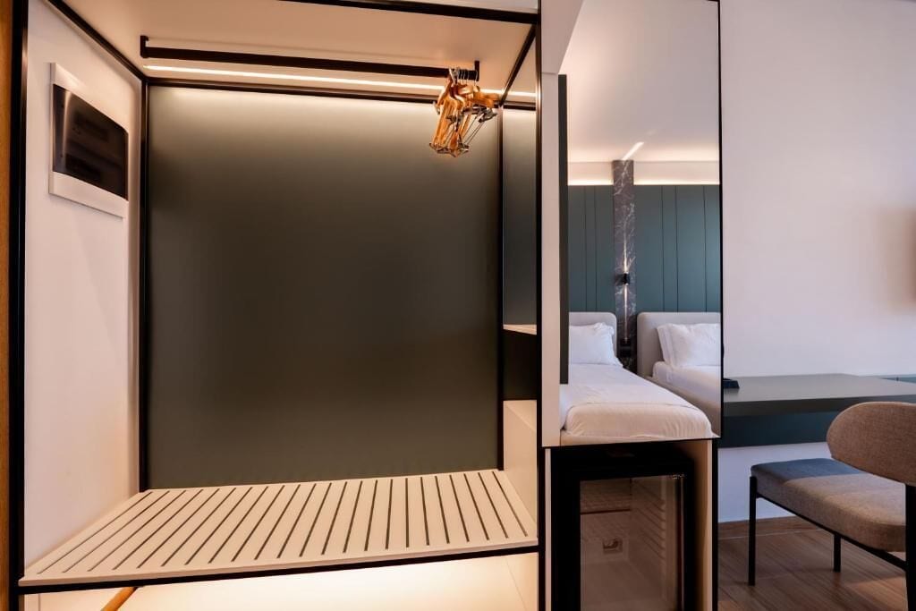 Egy modern hotelszoba, minimalista dizájnnal, amely egy ágyat, egy ülősarokot és egy egyedi, a falra szerelt lámpatestet tartalmaz.