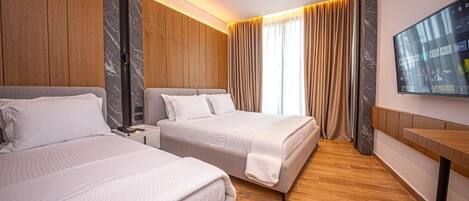 Una habitación de hotel con dos camas, suelo de madera, un televisor en la pared y un ventanal con cortinas.