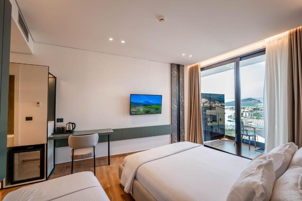 Habitación de hotel con cama, escritorio, silla y televisor. Amplia ventana con vistas a una ciudad y montañas.