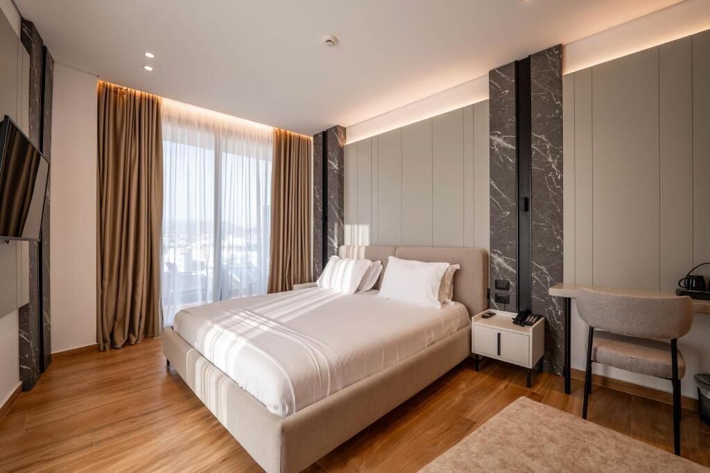 A modern hotel bedroom with a large bed, bedside tables, a chair, and a view of the city.