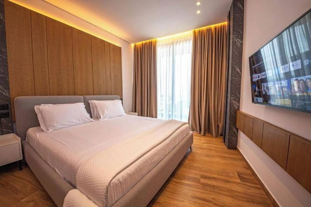 Una habitación de hotel moderna con una cama grande, paneles de madera, un televisor de pantalla plana y un amplio ventanal con cortinas.
