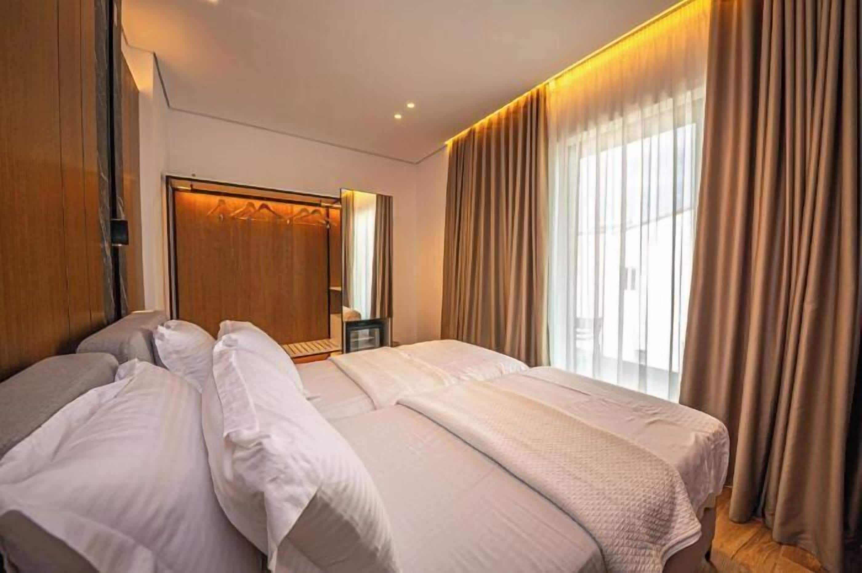A hotel room with two beds, a wooden wardrobe, a mirror, and a large window with curtains.