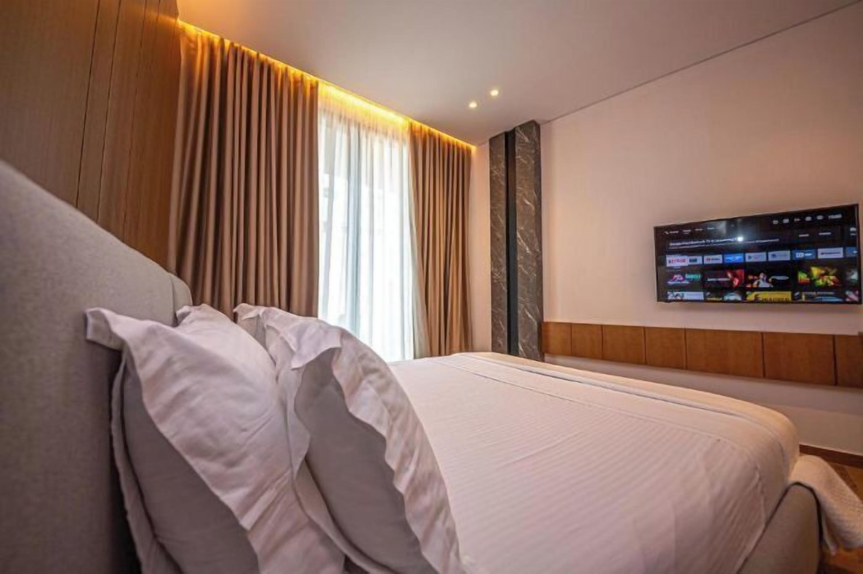 Ein Hotelzimmer mit Bett, Vorhängen, einem Flachbildfernseher an der Wand und einem hölzernen Kopfteil.