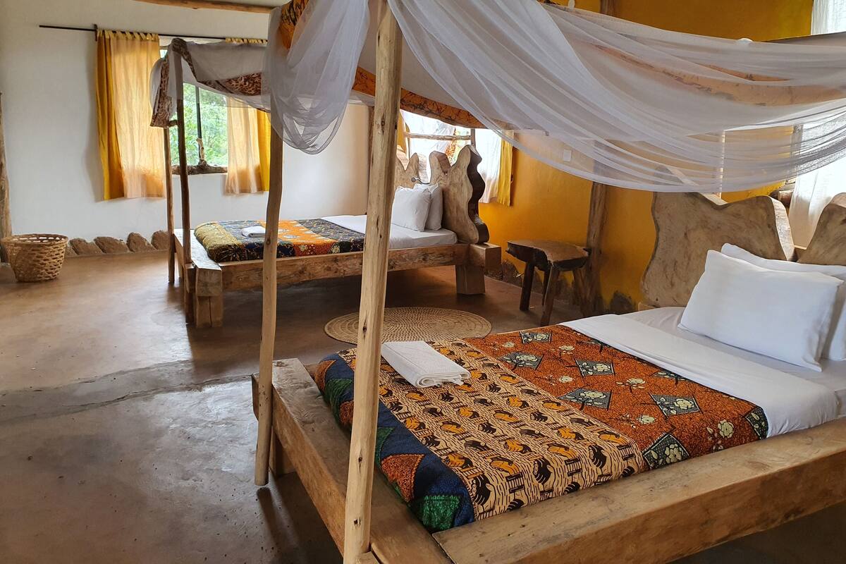 A rustic room with a bed under a canopy, another bed with a patterned bedspread, a woven basket, and a large wooden sculpture.