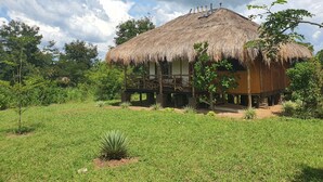 A thatched-roof house with a wooden deck, surrounded by lush greenery. - Topi Lodge (Kihihi)