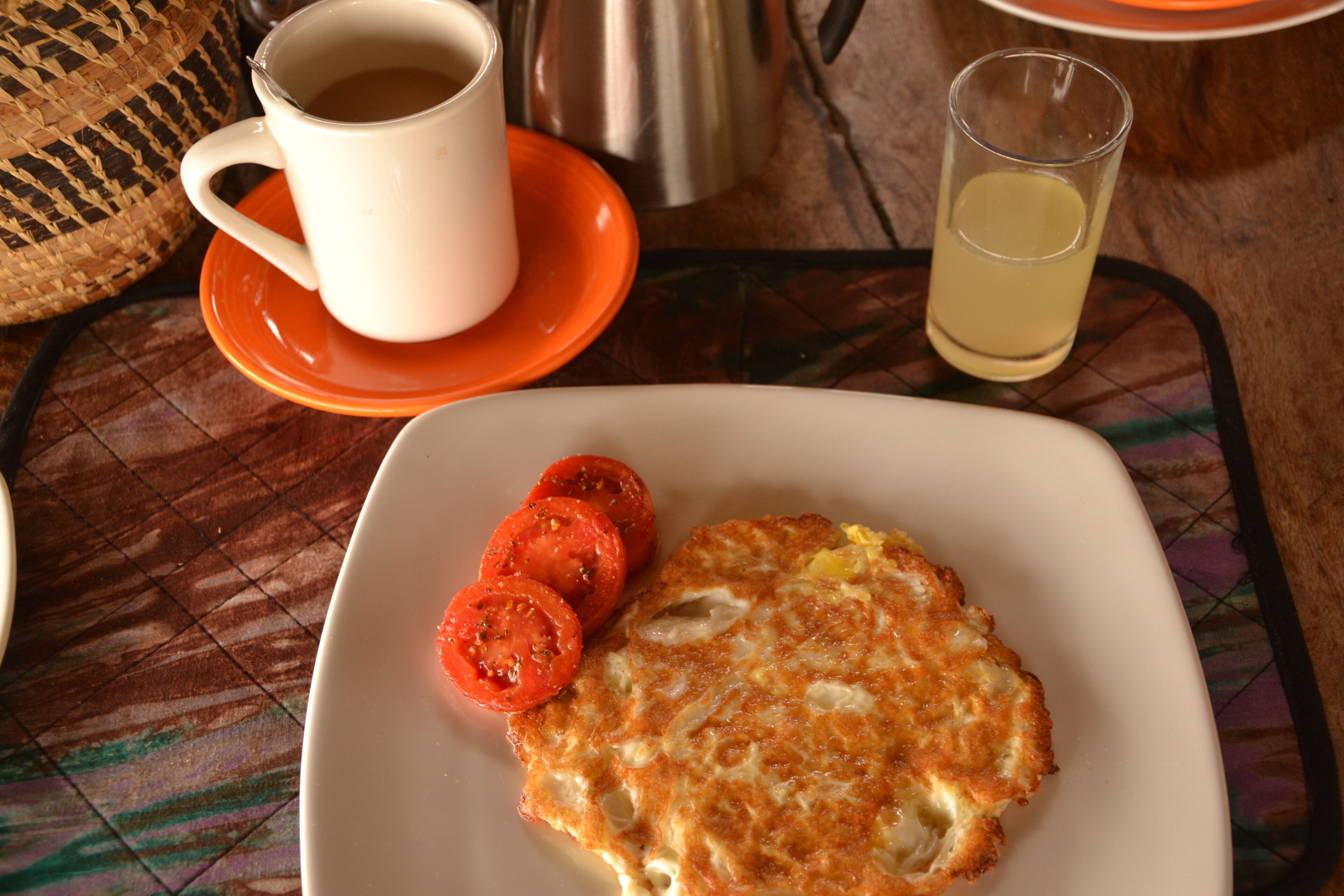 A plate with a fried egg and tomato slices, a white mug, and a glass of a yellowish liquid on a table with a woven basket and a metal pot in the background.