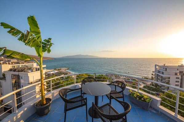 A terrace with a round table and chairs, a potted plant, and a view of the sea and distant land.