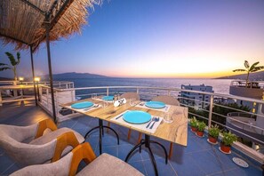 A dining table set for four with blue plates, surrounded by cushioned chairs, overlooking a coastal view at sunset.