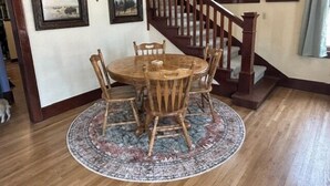 Un'area pranzo con un tavolo rotondo in legno e sedie, un tappeto a motivi, una scala con ringhiere in legno e quadri appesi alle pareti.