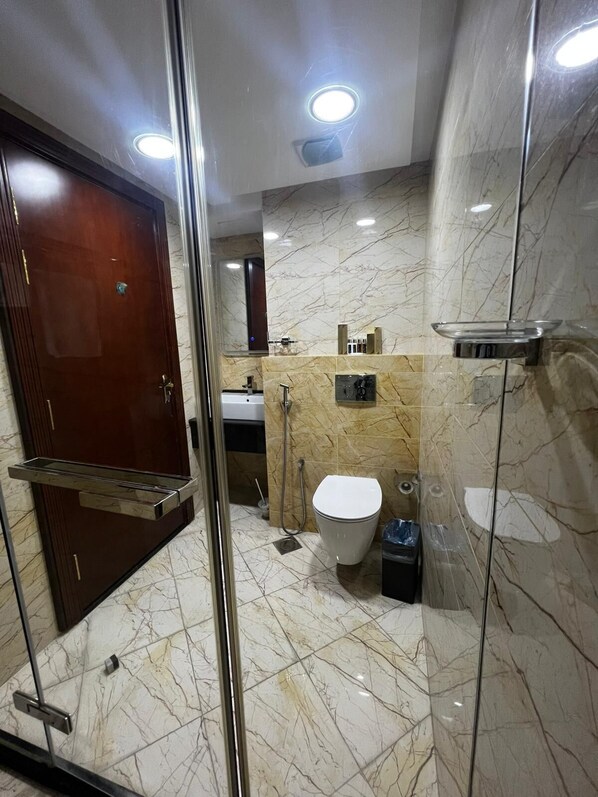 A modern bathroom with a glass shower enclosure, a marble-tiled floor, a white toilet, and a sink with a chrome faucet. - Saxophone Suites (Amwaj Islands)