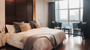 Ein modernes Hotelzimmer mit einem großen Bett, einer Sitzecke mit Sesseln, Blick auf die Stadt durch große Fenster und einer außergewöhnlichen Deckenleuchte.