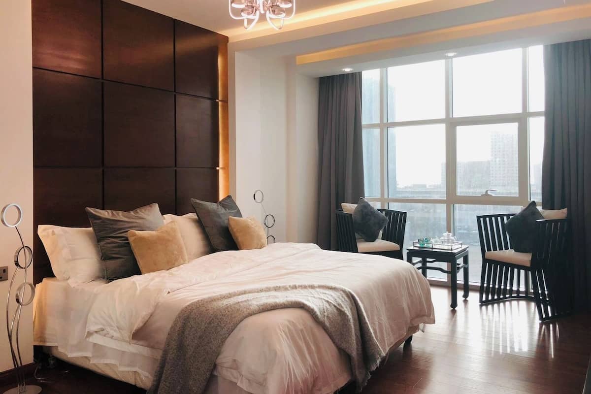A modern hotel room with a large bed, a seating area with chairs, a view of the city through large windows, and a unique ceiling light fixture.