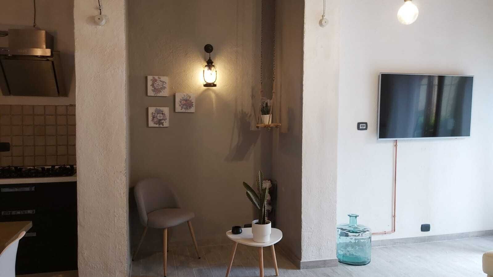A modern living room with a flat-screen TV, a small table with a potted plant, a grey chair, and a wall-mounted light fixture.