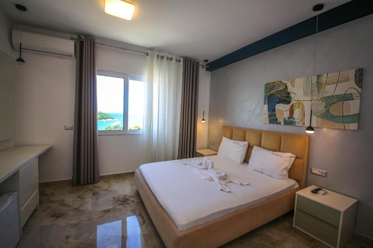 A hotel room with a bed, bedside tables, a window with curtains, and a painting on the wall.