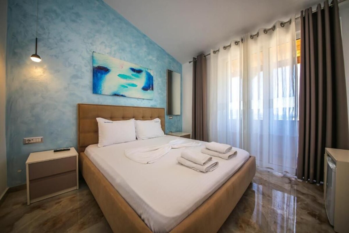 A hotel room with a large bed, a nightstand, a painting on the wall, and a window with curtains.