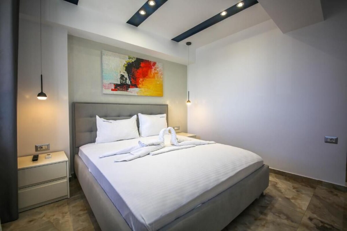 A modern hotel room with a large bed, a nightstand, a painting on the wall, and recessed ceiling lighting.