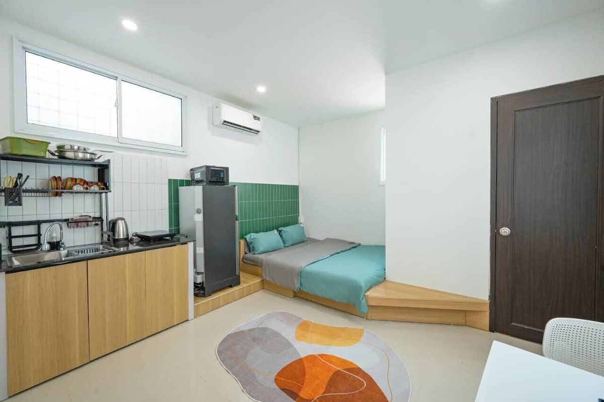 Ein kleines Zimmer mit Küchenzeile, einem Bett mit grünem Kopfteil, einer Holztür und einem weißen Stuhl.