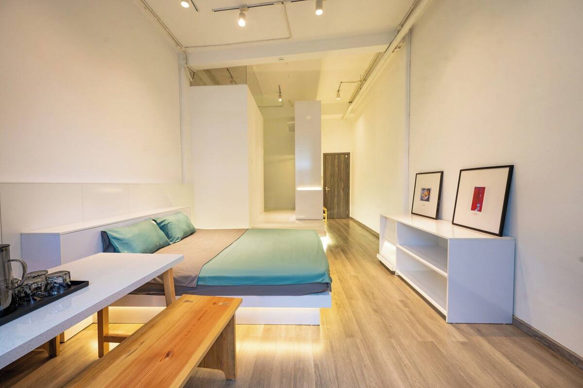 Ein moderner, minimalistischer Raum mit Bett, Sitzbank, kleinem Tisch und Regal mit gerahmten Bildern.