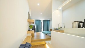 Habitació d'hotel moderna amb sòl de fusta, un llit amb capçalera blava, una petita taula d'escriptori amb una làmpada, una planta i una papereta a la barra.
