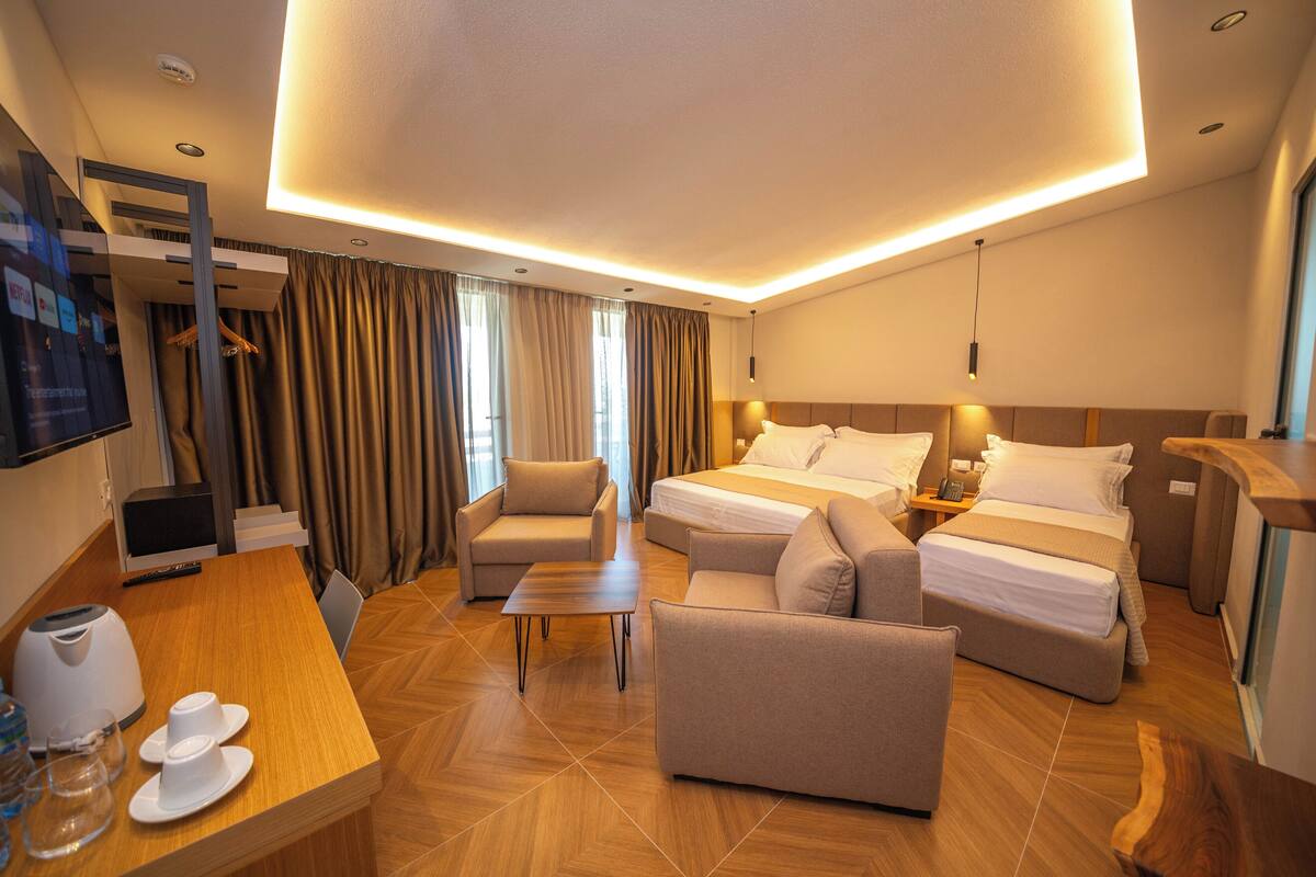 A hotel room with a bed, two sofas, a coffee table, a desk with a kettle and cups, and a TV.