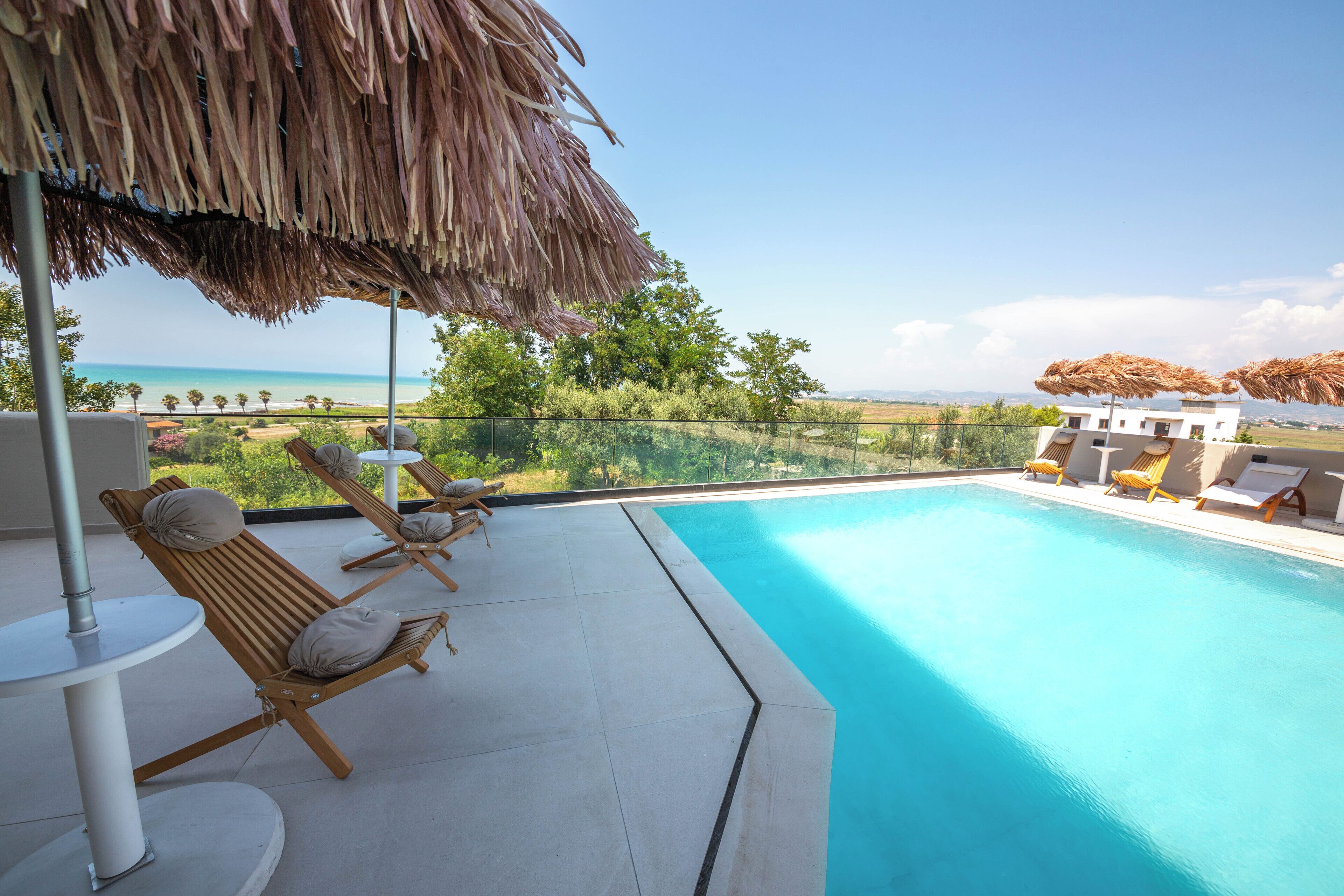 Un espace aménagé au bord de la piscine, avec des chaises longues et un parasol en chaume, offrant une vue sur une piscine aux eaux cristallines et une plage lointaine.
