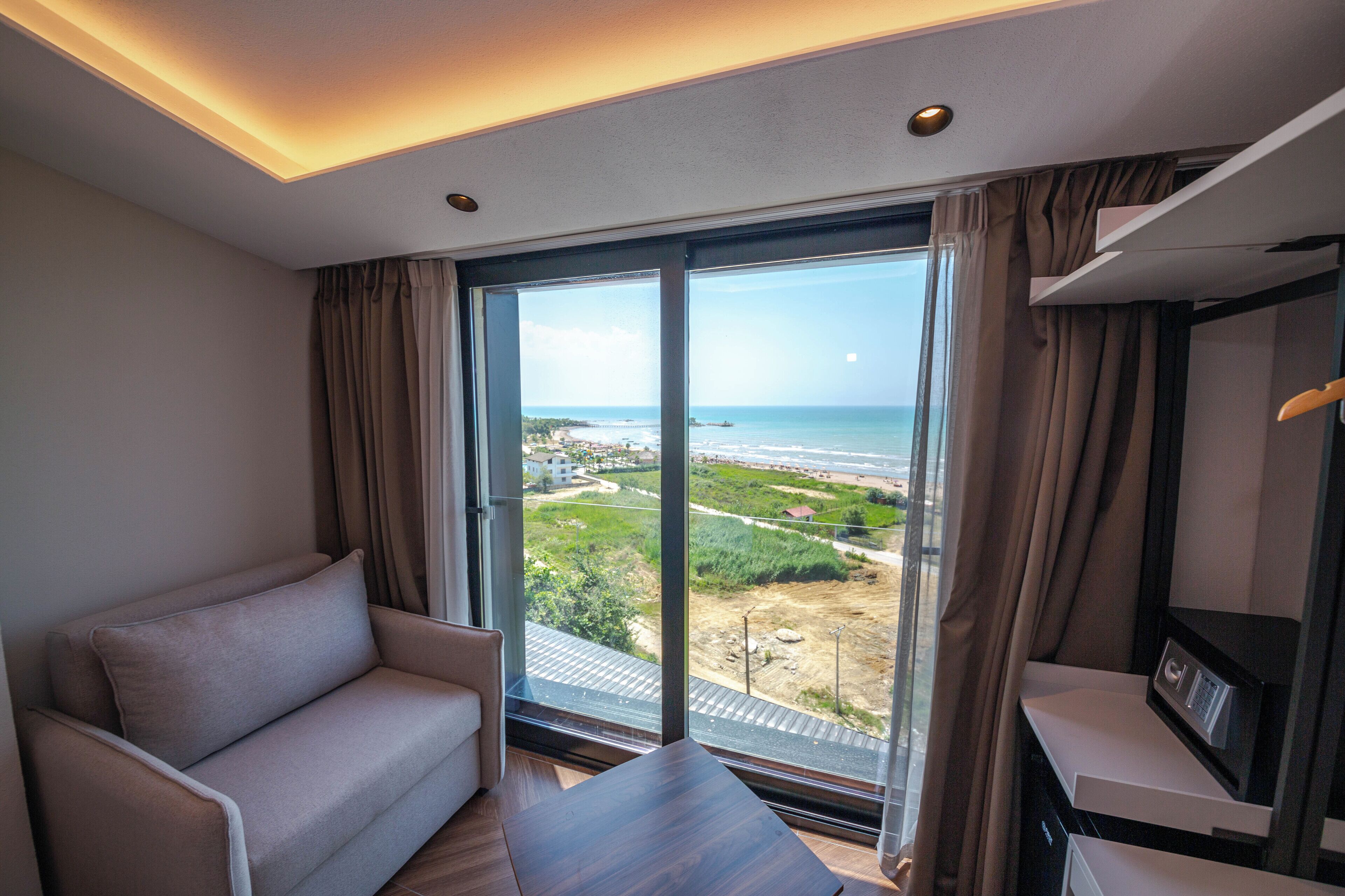 Une chambre d’hôtel moderne dotée d’une grande porte-fenêtre coulissante donnant sur un balcon avec vue sur l’océan et une côte lointaine.