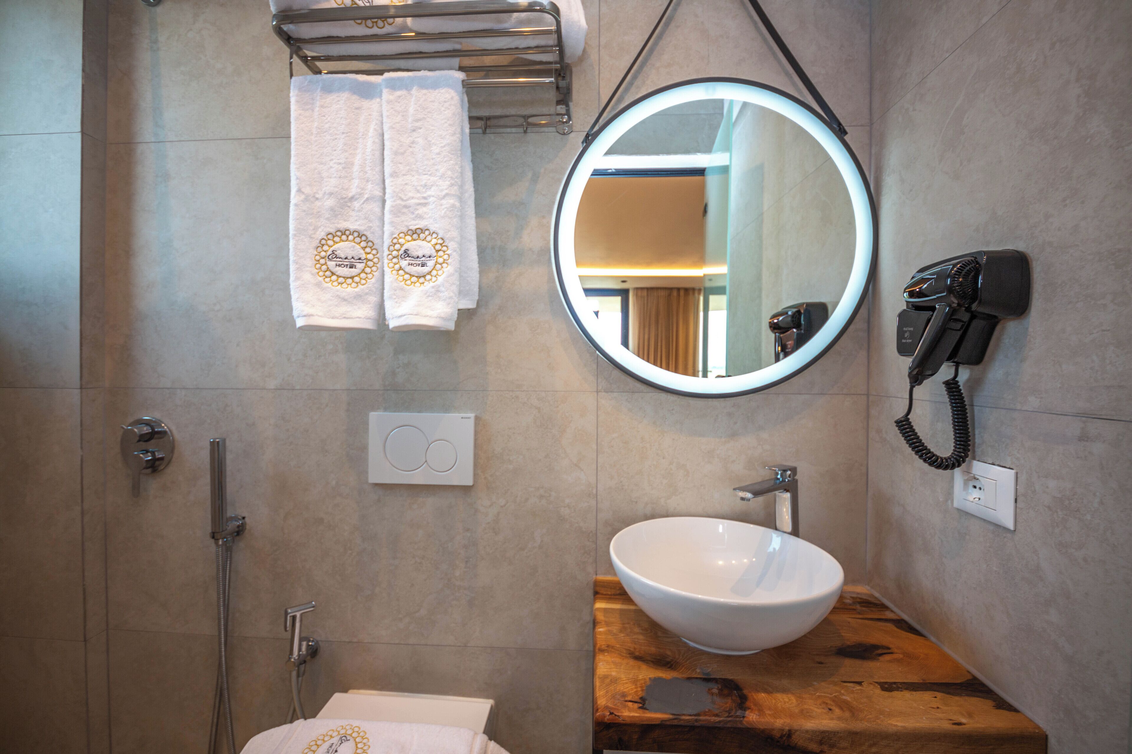 Un baño con tocador de madera, un lavabo blanco, un espejo redondo, un perchero con dos toallas, un secador de pelo portátil y una ducha con cabezal de ducha manual.