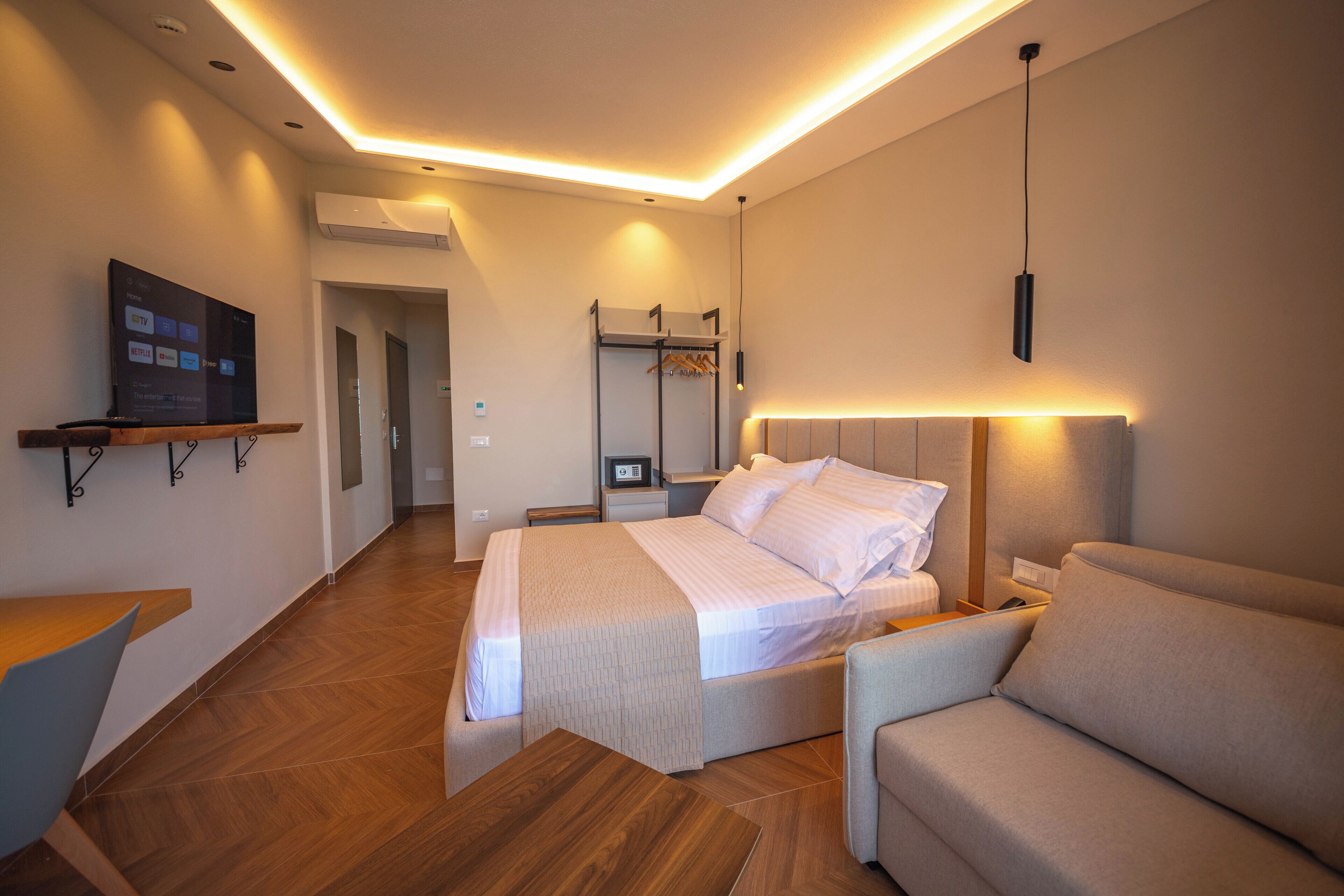 A modern hotel room with a bed, a sofa, a TV on a stand, and a coat rack.