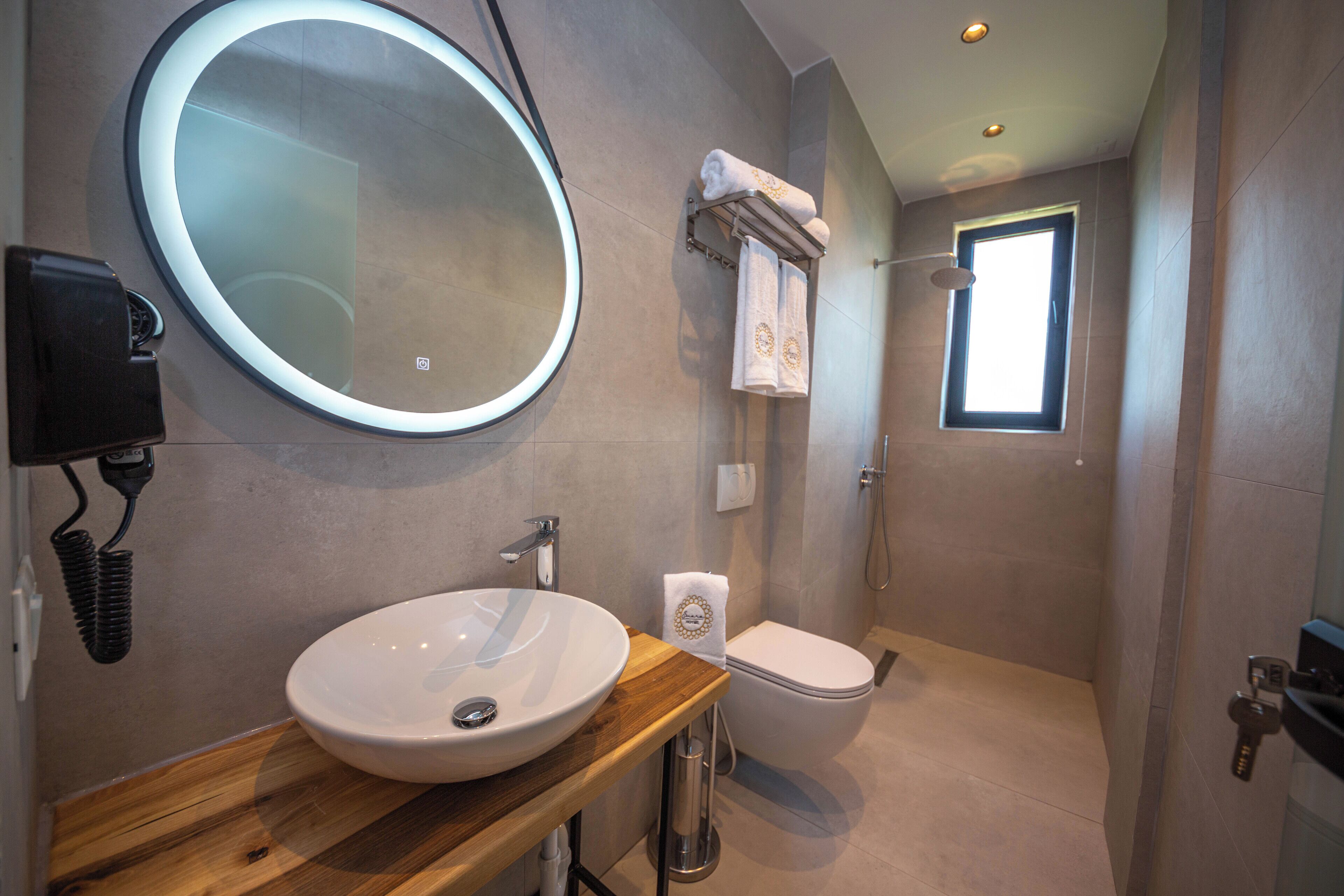 A modern bathroom with a round mirror, a wooden vanity, a white sink, a toilet, and a shower area.