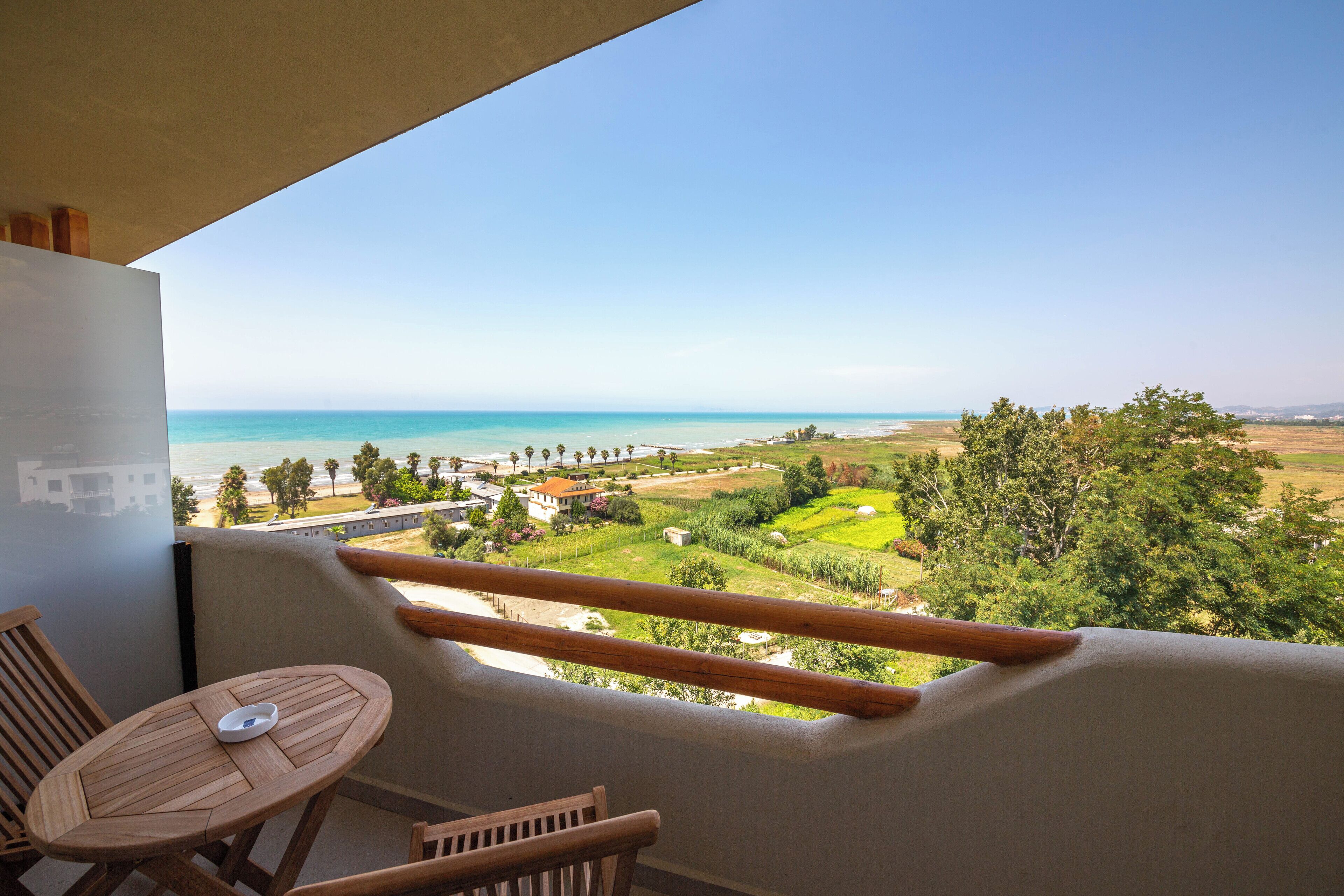 Un balcón con mesa y sillas de madera, con vista a un paisaje costero y una vista despejada del mar y edificios a lo lejos.